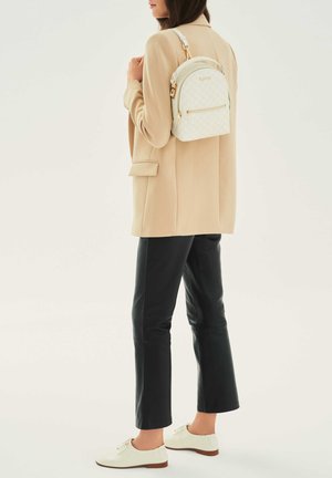 Zaino bianco trapuntato con hardware dorato, design arrotondato e manico superiore. Indossato sopra un blazer beige e pantaloni di pelle nera con scarpe bianche.