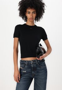 Črn skrajšan pleten majica s kratkimi rokavi, ki ima majhen logotip okoli ovratnika, v kombinaciji z raztrganimi modrimi jeans hlačami in sijočo črno torbico.