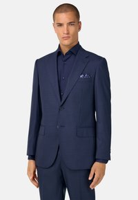 Traje azul marino a medida con patrón de rayas, cierre de un solo botón, solapas de pico y bolsillos laterales. Complementado con una camisa a juego y un pañuelo de bolsillo.