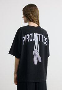 Γυναίκα με μακριά καστανά μαλλιά που φοράει μαύρο μπλουζάκι με εικόνα παπουτσιών μπαλέτου και τη λέξη "PIROUETTES" στην πλάτη, σε απλό φόντο.