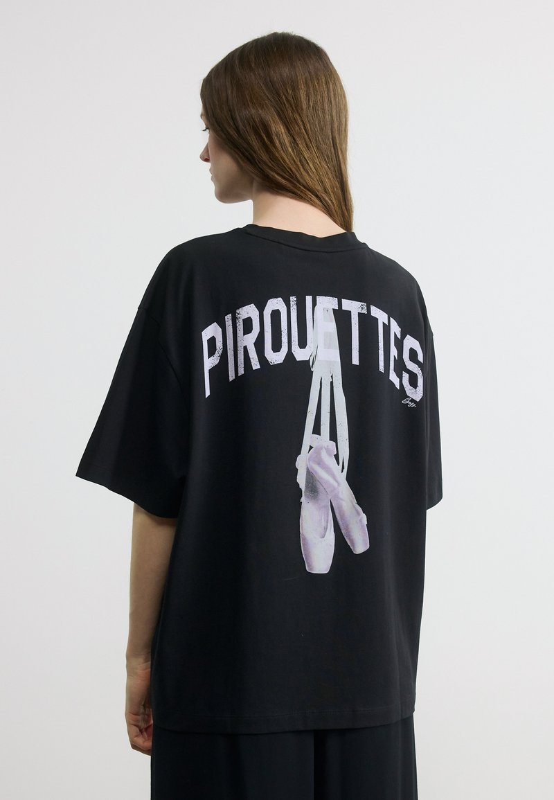 Γυναίκα με μακριά καστανά μαλλιά που φοράει μαύρο μπλουζάκι με εικόνα παπουτσιών μπαλέτου και τη λέξη "PIROUETTES" στην πλάτη, σε απλό φόντο.