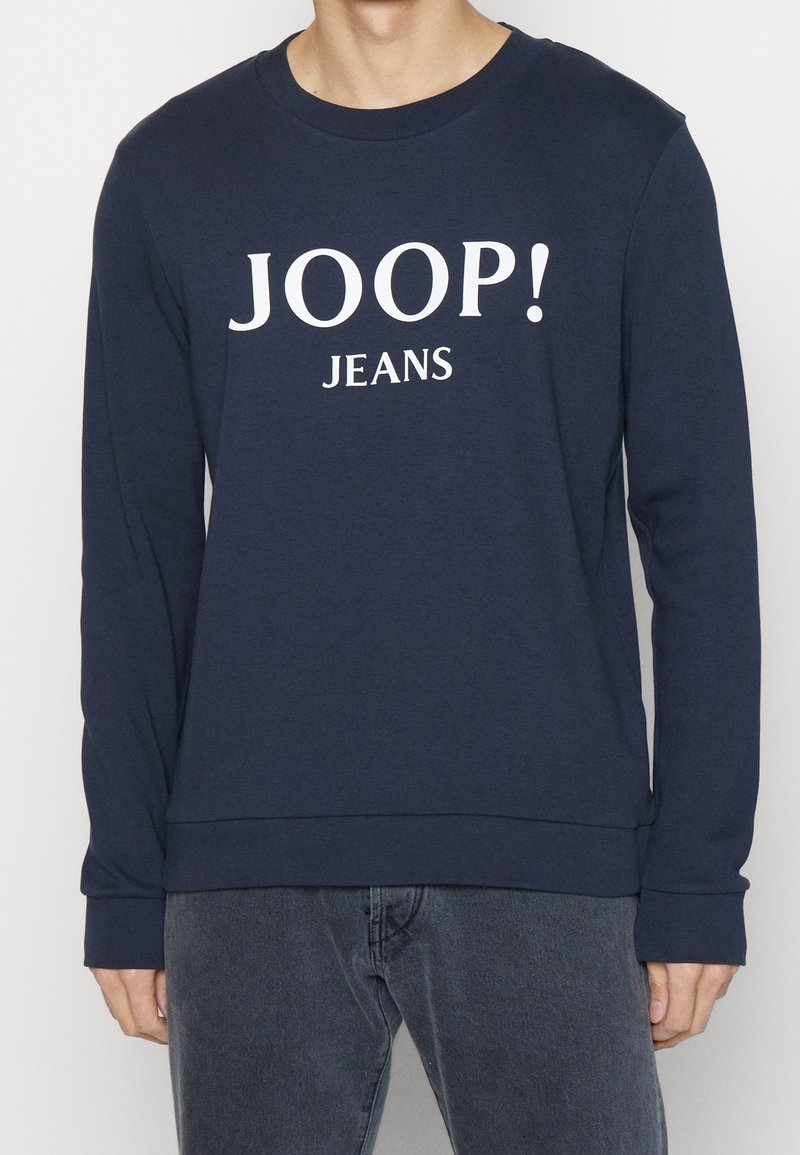 Homme portant une chemise bleu marine à manches longues avec le texte blanc « JOOP! JEANS », associée à un jean gris foncé, debout devant un fond blanc.
