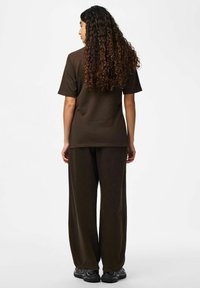 Femme aux longs cheveux bouclés portant un t-shirt à manches courtes marron foncé et un pantalon ample, debout de dos sur fond blanc.