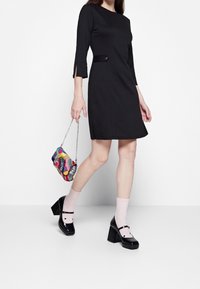 Musta polvipituinen mekko, jossa on 3/4-hihat ja sivuhalkaisu, yhdistetty moniväriseen käsilaukkuun, vaaleanpunaisiin sukkiin ja mustiin lohkopohjakenkiin.