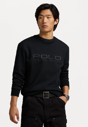 Melnā sporta krekls ar "POLO RALPH LAUREN" logotipu reljefa tekstūrā. Arapa kakla, garām piedurknēm, uzvilkts pār melnajām biksēm ar jostu.