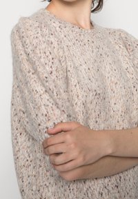 Gruby sweter w jasnym beżu z brązowymi i pomarańczowymi wstawkami. Okrągły dekolt, krótkie rękawy i luźny krój. Miękka faktura.