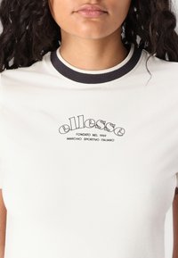 Balta t-krekls ar īsām piedurknēm un apaļu melnu ribota kakla izgriezumu. Ar melnu logo druku "ellesse" un papildu tekstu apakšā.