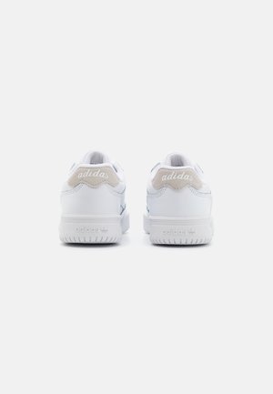 Para białych tenisówek Adidas widocznych od tyłu, z beżowymi zamszowymi wstawkami na pięcie z białym napisem logo Adidas.