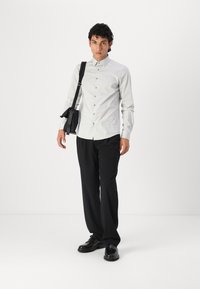 Lichtgrijs overhemdbed met een spreadcollar, zwarte pantalon en zwarte schoenen. Model draagt een zwarte schoudertas.