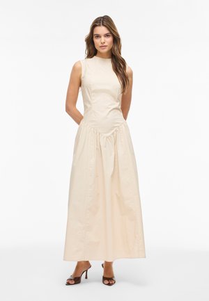 Pikkade pruunide juuste ja varrukateta beežika maksi-kleidiga naine, kes kannab pruune avatud varvastega kontsakingi, seisab käed selja taga.