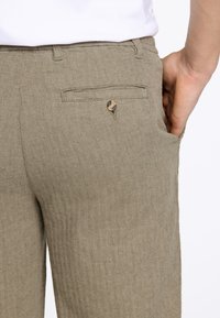 Kakhihosen mit Fischgrätenmuster, die eine Gesäßtasche mit Knopfverschluss haben; der Stoff hat eine strukturierte, gewebte Optik.