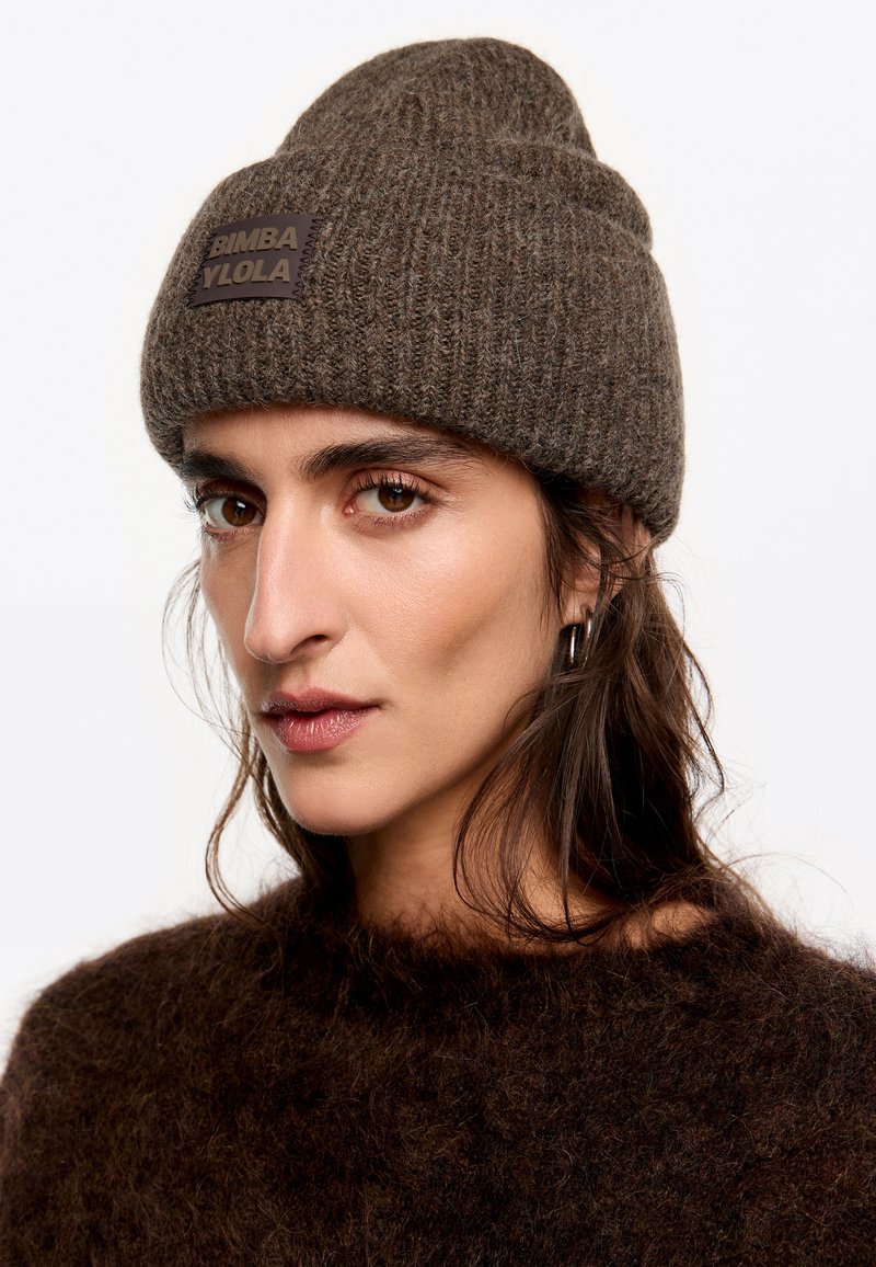 Brązowa czapka dzianinowa z prążkowaną fakturą i wyraźną naszywką marki, stylizowana z podwiniętym mankietem. Miękki materiał, casualowy design.