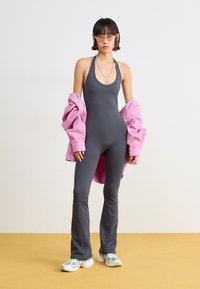 Grå jumpsuit med en tettsittende design, kombinert med en løs rosa kordjakke. Fullført med sportslige sko som har en blanding av farger og teksturer.