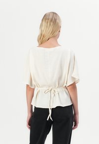 Blusa color crema realizzata in un tessuto leggero con una vestibilità ampia, maniche larghe e un dettaglio di vita annodato. La vista posteriore mostra un design semplice.