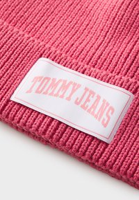 Růžová pletená čepice s ribovanou texturou. Obsahuje obdélníkovou bílou etiketu s nápisem "TOMMY JEANS" v světle růžovém písmu.