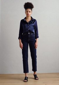 Navy Satin Hemd mit langen Ärmeln, kombiniert mit hochgeschnittenen, dunklen Jeans und schwarzen, offenen Plataforma-Pumps.