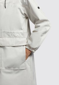 Giacca leggera color crema con una comoda tasca frontale, polsini elasticizzati e un discreto logo sul petto. Tessuto liscio e resistente all'acqua.