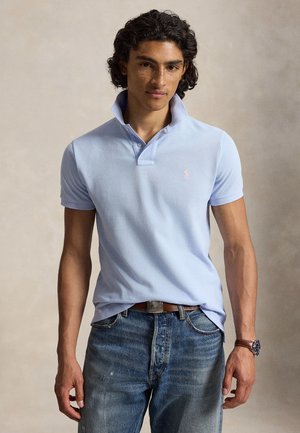 Młody mężczyzna z kręconymi włosami, ubrany w jasnoniebieską koszulkę polo, niebieskie dżinsy, brązowy pasek i zegarek na rękę, stojący na jednolitym tle.