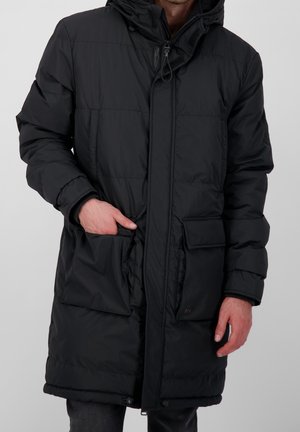 Man met een lange zwarte gewatteerde winterjas met capuchon, handen bij twee grote voorzakken, staand tegen een eenvoudige achtergrond.