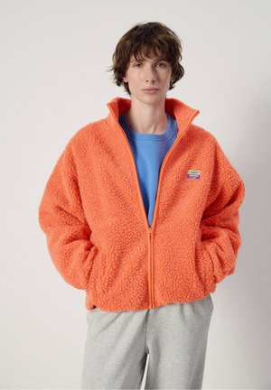 Personne aux cheveux bruns courts portant une veste polaire orange par-dessus une chemise bleue et un pantalon gris clair, debout devant un fond uni.