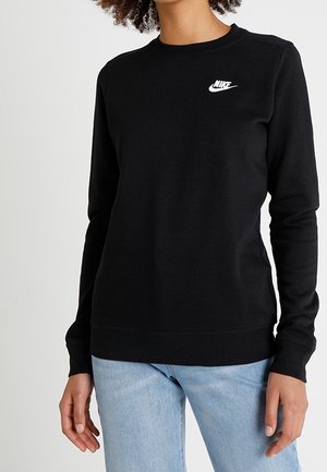 Kobieta ubrana w czarną bluzę Nike z białym logo na piersi i jasnoniebieskie dżinsy, stojąca z rękami luźno opuszczonymi wzdłuż boków.