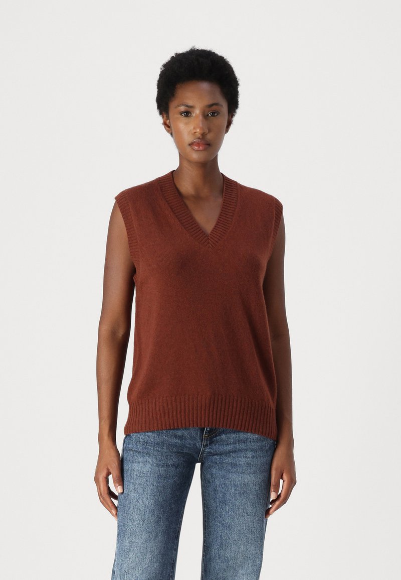 Leinenfarbener ärmelloser Pullover mit V-Ausschnitt, geripptem Saum und subtiler Textur. Kombiniert mit blauen Jeans.