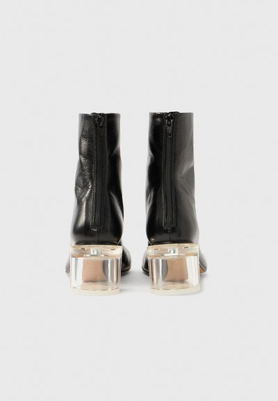 Černé kožené kotníkové boty s hladkou texturou, bočními zipy a průhlednými blokovými podpatky. Design se vyznačuje minimalistickým, elegantním tvarem.
