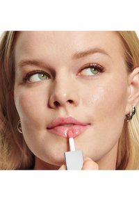 Läppglans i fyrkantig förpackning med en vit applikator, som appliceras på mjuka, glansiga rosa läppar. Modellen har en jämn hy med naturliga tonfall.