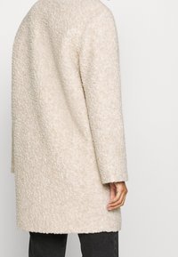 Person med en cremede tekstureret uldfrakke, der står med den ene hånd afslappet ved siden af, iført mørke bukser.