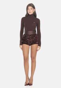 Burgunderfarbenes geripptes Rollkragenoberteil mit einer kleinen Tasche, kombiniert mit glitzernden hochgeschnittenen Shorts. Das Model trägt passende burgunderfarbene Absätze.