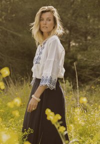 Witte geborduurde blouse met blauwe accenten, 3/4 bell mouwen; marineblauwe lange rok; lichte armband en ringaccessoires, omringd door gele bloemen.