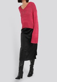 Rosa Strickpullover mit V-Ausschnitt und langen Ärmeln, kombiniert mit einem schwarzen Satin-Rock und schwarzen, kniehohen Absatzstiefeln.