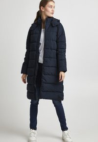 Manteau matelassé bleu marine avec capuche, doté d'une fermeture éclair et d'une texture matelassée. Porté par-dessus un haut blanc et associé à un jean foncé et des baskets blanches.