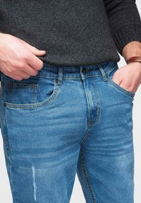 Jean en denim bleu clair à coupe slim, avec des poches avant, des détails de décoloration et des coutures contrastantes. Porté avec un pull foncé.