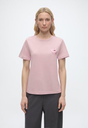 Blondynka w prostej jasnoróżowej koszulce z małym kwiatowym wzorem na lewej piersi, w połączeniu z ciemnoszarymi spodniami.