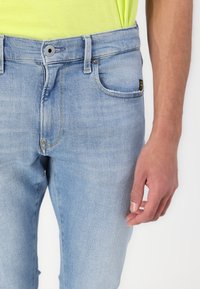 Világoskék farmernadrág, slim fit szabással, ötpocket dizájnnal, gombos zárással, finom varrási részletekkel. A textúrált anyag jól látható.