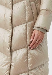 Beige gewatteerde jas met een doorgestikt patroon, gemaakt van een gladde, glanzende stof. Bevat een gebreide manchet aan de mouw en een hand die op de stof rust.