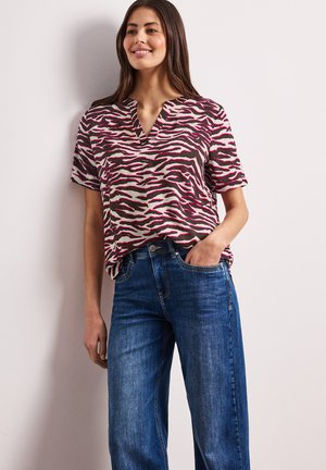 Kobieta ubrana w beżowo-różowy top w zebra print z krótkim rękawem, włożony w wysokie, niebieskie dżinsy, stojąca z jedną ręką w kieszeni.