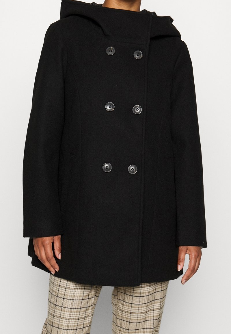 Manteau en laine noir à double boutonnage avec capuche, six boutons, porté sur un pantalon beige à carreaux, montrant le haut du corps et les mains.