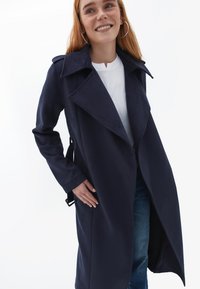 Trench coat navy hecho de una tela suave; cuenta con solapas anchas, un cinturón atado en la cintura y bolsillos laterales. Lleva encima una blusa blanca.