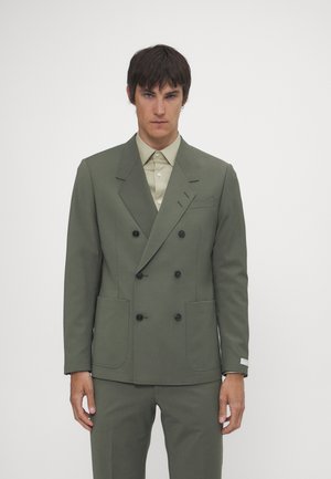 Dubbelgebreid blazer in olijfgroene stof, met twee voorkruiszakken, zwarte knopen en een lichtgroene overhemd met kraag eronder.