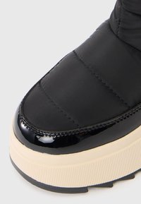 Zwarte gewatteerde sneaker met een glanzende zwarte neus, crèmekleurige tussenzool met ribbelstructuur en een textuur op de buitenstof.