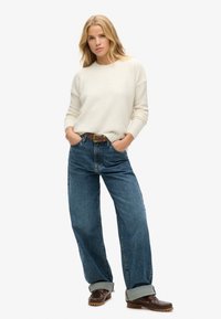 Kremowy sweter uszyty z miękkiego materiału, w połączeniu z luźnymi, niebieskimi dżinsami o wysokim stanie i podwiniętymi mankietami, dopełnionymi brązowymi butami.