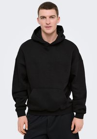 Jeune homme portant un sweat à capuche noir uni et oversize avec poche avant, debout devant un fond clair uni.