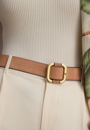 Cinturón de ante marrón claro con hebilla cuadrada dorada, llevado sobre un top de canalé crema y pantalones beige, parte de una manga floral verde y beige visible.