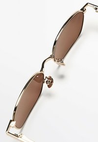 Occhiali da sole metallici con forma ovale, montatura dorata e lenti color marrone. Presentano un ponte nasale decorativo e accenti sottili.