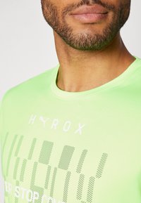 Camiseta deportiva de color verde neón, hecha de tela suave, con el texto "HYROX" y rayas estampadas en blanco. Cuello redondo y mangas cortas.