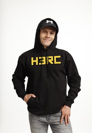 Mężczyzna w czarnej bluzie z kapturem z żółtym napisem "H3RC", czarnej czapce z białym logo i wyblakłych dżinsach, uśmiechający się z jedną ręką w kieszeni.