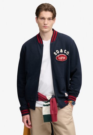 Giovane uomo che indossa una giacca blu navy con strisce rosse, una camicia bianca, pantaloni beige, un maglione annodato e un orologio da polso.