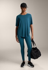 Türkiz edzés póló és leggings, fekete sportcipővel párosítva. A modell egy fekete sporttáskát tart. Simább textúra, laza fazon, elegáns dizájn.